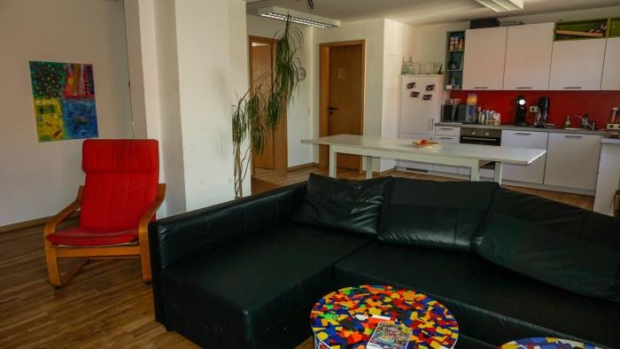 Wohnzimmer mit schwarzer Ledercouch, Küchenzeile im Hintergrund und bunten Beistelltischen Wohnzimmer mit schwarzer Ledercouch, Küchenzeile im Hintergrund und bunten Beistelltischen