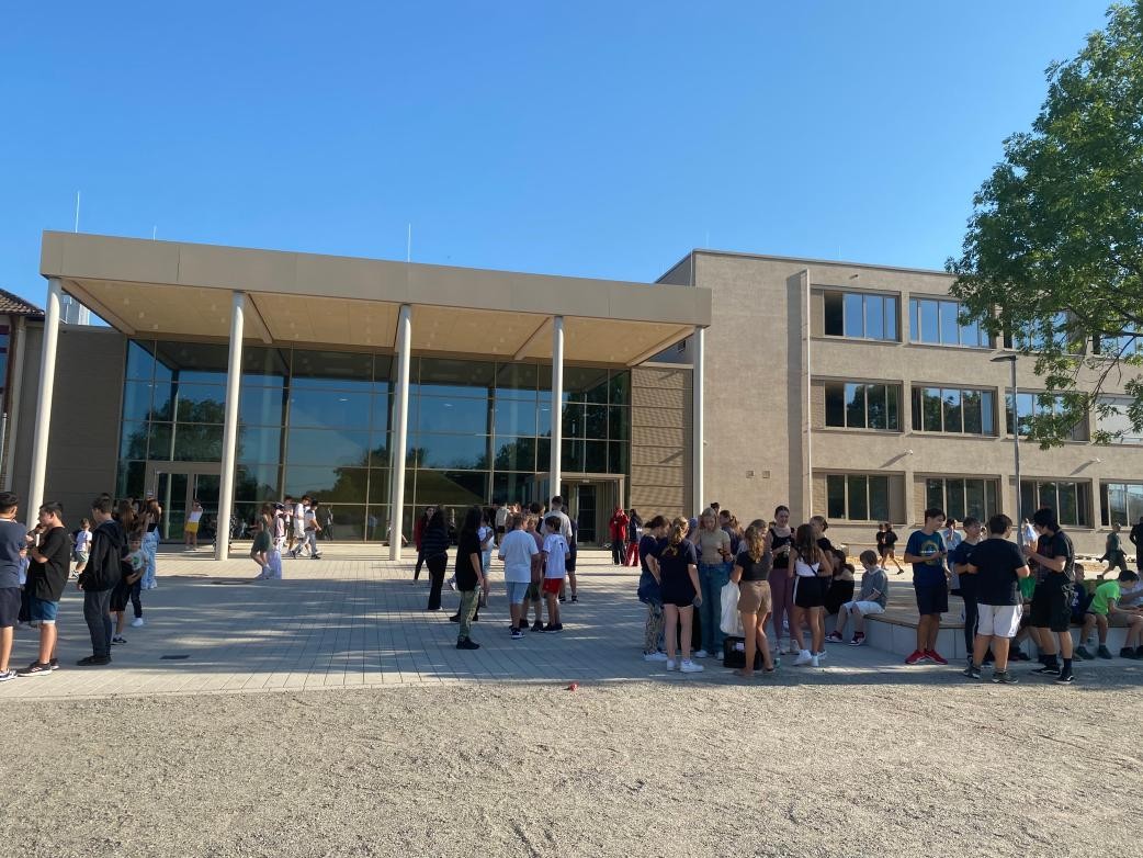 Verschiedene Gruppen von Menschen stehen vor der Fassade des Schulneubaus mit großer Glasfassade, einem vorspringenden Vordach, getragen durch schlanke Säulen